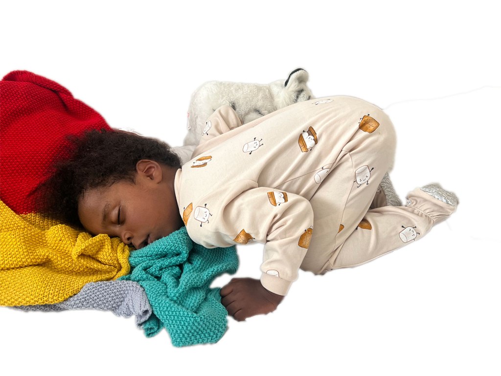 A 2 year old black boy is sleeping in a cream-coloured onesie with smores and marshmallows dancing on them. the right side of his face is down, he has a teddy tucked on his right side, his knees are tucked to his tummy and his bottom is in the air. his left hand is holding the edge of a handknit blanket, knit in a moss stich in green, grey, red, and yellow.