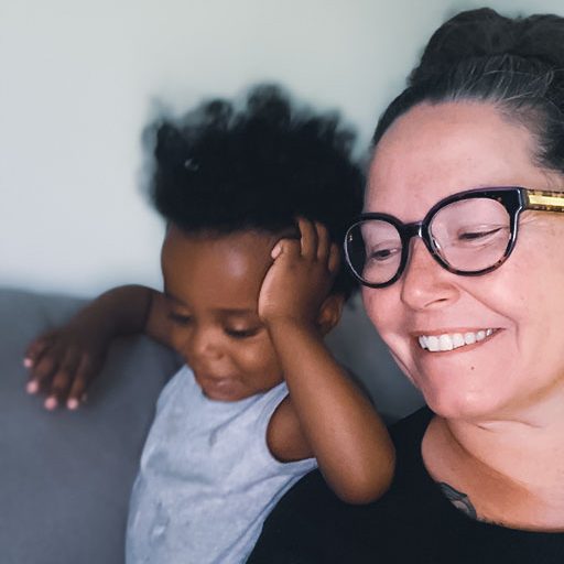a 2year old black toddler wearing a grey sleeveless onesie is leaning on his grandmothers right shoulder with his left elbow, his left hand resting on his face. His grandmother is a Caucasian woman of an indeterminate age, wearing a black T-shirt, dark purple plastic-framed glasses, and her salt-and-pepper hair is up on top of her head. The tow are sharing a happy moment on a grey sofa; it looks like they are reading a book.