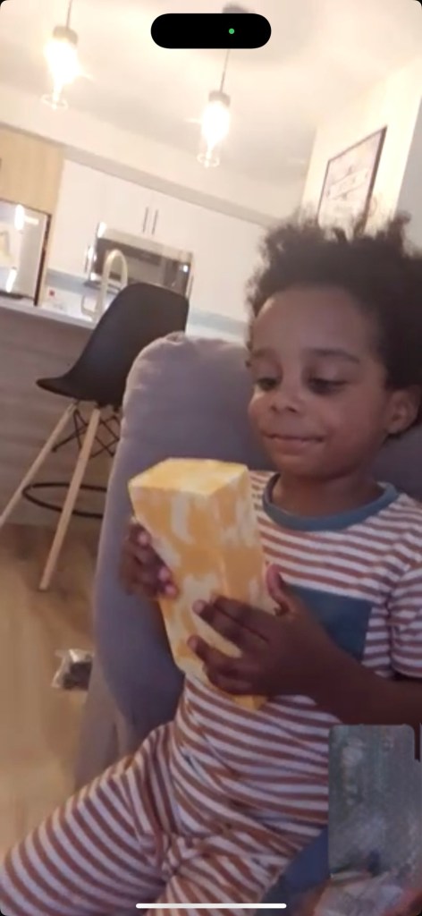 A 3 year old black boy in red & white stripped pyjamas gazes down cheekily at a Costco-sized brick of marbled cheese that he is holding with both hands in front of him. He is sat on the edge of a grey sofa with the view of a kitchen in the background. 