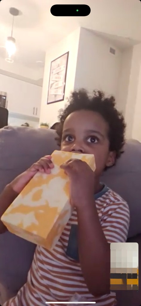 A 3 year old black boy in red & white stripped pyjamas chomps down on a Costco-sized brick of marbled cheese that he is holding with both hands in front of him. He is sat on the edge of a grey sofa with the view of a kitchen in the background. 