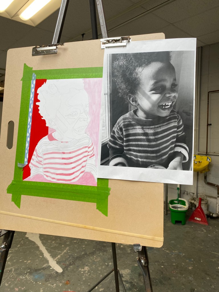 An in-progress painting of a young black boy, Ace, on a brown drawing board in a studio setting. The painting is taped down with green tape and a reference black-and-white photo is clipped beside it. The portrait shows Ace smiling, with the face still being developed using grayscale watercolour. The striped shirt is outlined and partially filled in with pink and red tones. A red acrylic background fills the left side of the composition, while the right remains pale pink.