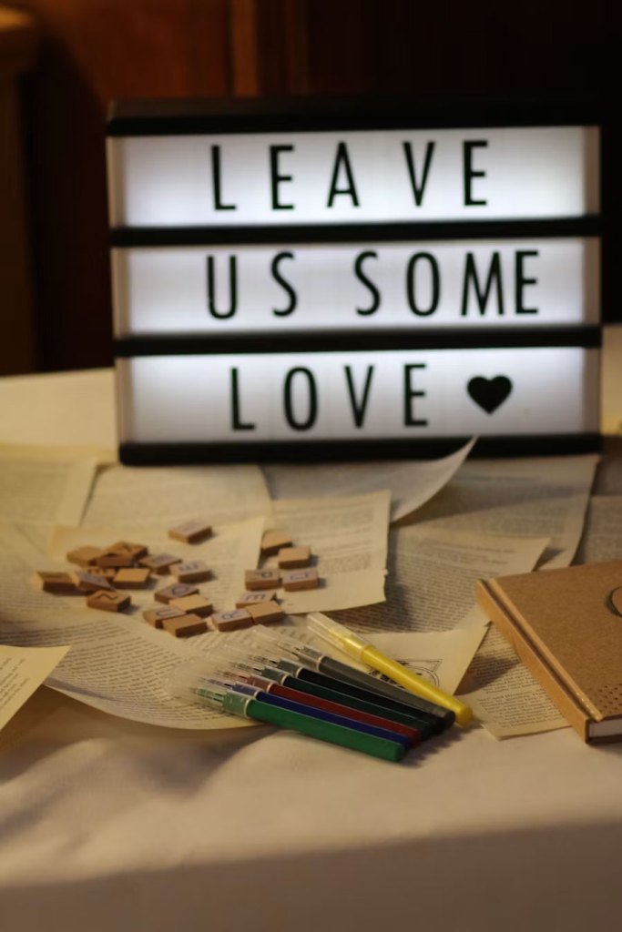 A warm, softly lit tabletop display featuring a light-up sign that reads “LEAVE US SOME LOVE” in black capital letters on a white background, with a black heart icon beneath the text. Surrounding the sign are scattered, loose book pages creating a textured paper backdrop. Scrabble-style wooden letter tiles and a row of colorful gel pens lie across the pages, inviting interaction. A tan, hardcover notebook sits at the edge, completing the cozy, craft-inspired setup — perfect for a guestbook or creative message space.