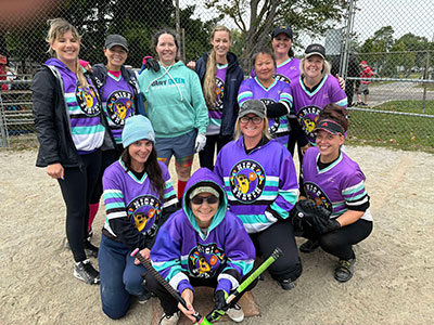 Eleven ladies wearing purple and minty green baseball jerseys. may are wearig coats and hats so it was a chilly day when the summer softball season was over, they are grouped together, smiling.
