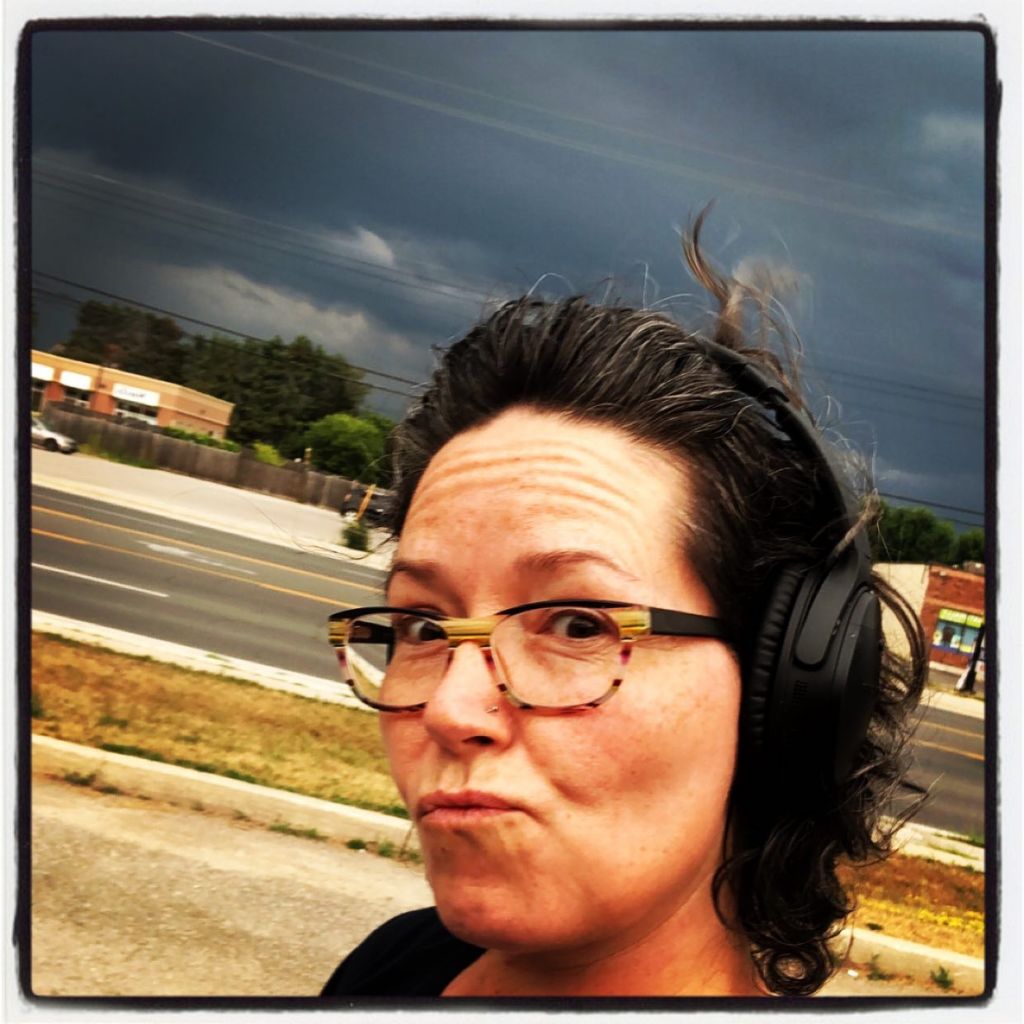 The sky in the background is dark and threatening a storm. A Caucasian woman with short dark hair is walking on a town street, wearing black Bose headphones and dark-ish glasses frames. Her mouth is scrunched in an exagerrated gesture of questioning the impending weather.