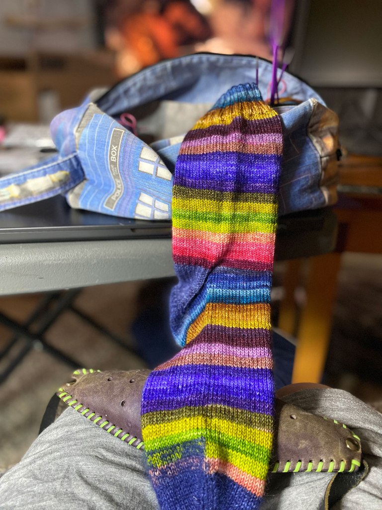 A hand knit sock in blended stripes of deep, rich jewellery tones of purple, blue, magenta, with shots of lime green, lilac, and mustard to make it mesmerizing. it is draped against a knitting bag in the pattern of the TARDIS. THere is a knitting belt under the sock and on the lap of the knitter.