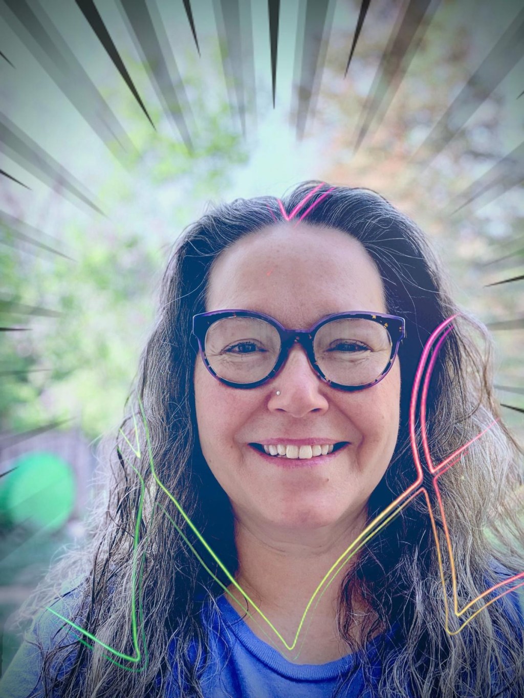 A smiling Caucasian woman with long, dark wavy gray-streaked hair and dark purple tortoiseshell glasses poses outdoors. She’s wearing a blue shirt and standing in soft sunlight with a blurred background of trees and a fence. The image is edited with Photoshop Express to include bold comic-style burst lines radiating from her head, and a rainbow neon streak that implies a heart shape framing her faceadding a dramatic, energetic flair.