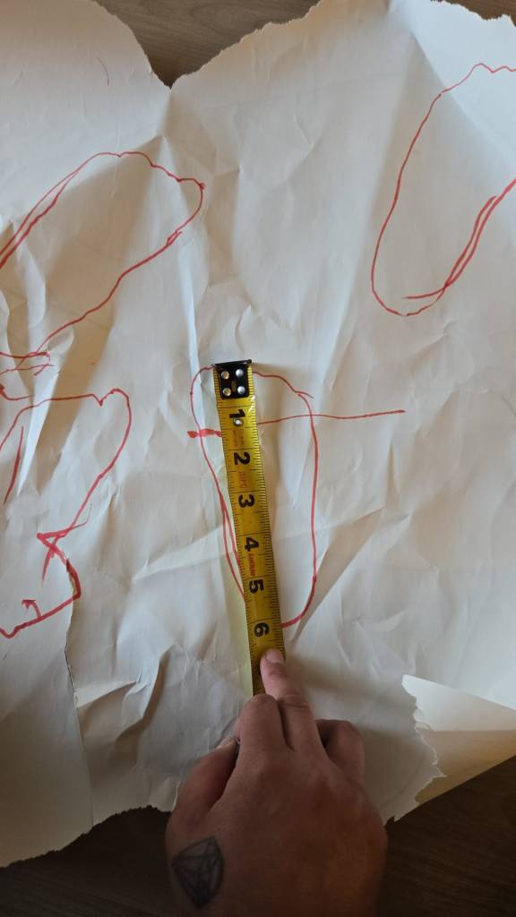 A crinkled piece of paper with multiple red foot outlines drawn on it. A traditional yellow measuring tape stands vertically over one tracing, showing a foot length of approximately 6 inches. An adult’s hand holds the tape measure in place.