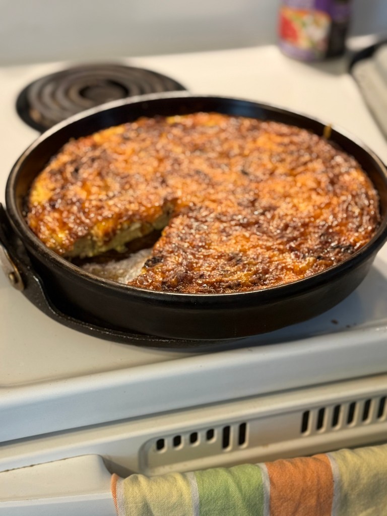 A golden, cheesy baked egg and cottage cheese dish in a black cast iron skillet. A wedge has been cut out to reveal the soft, textured interior filled with caramelized onions. The pan sits on a white stovetop with a multicoloured tea towel draped over the oven handle.