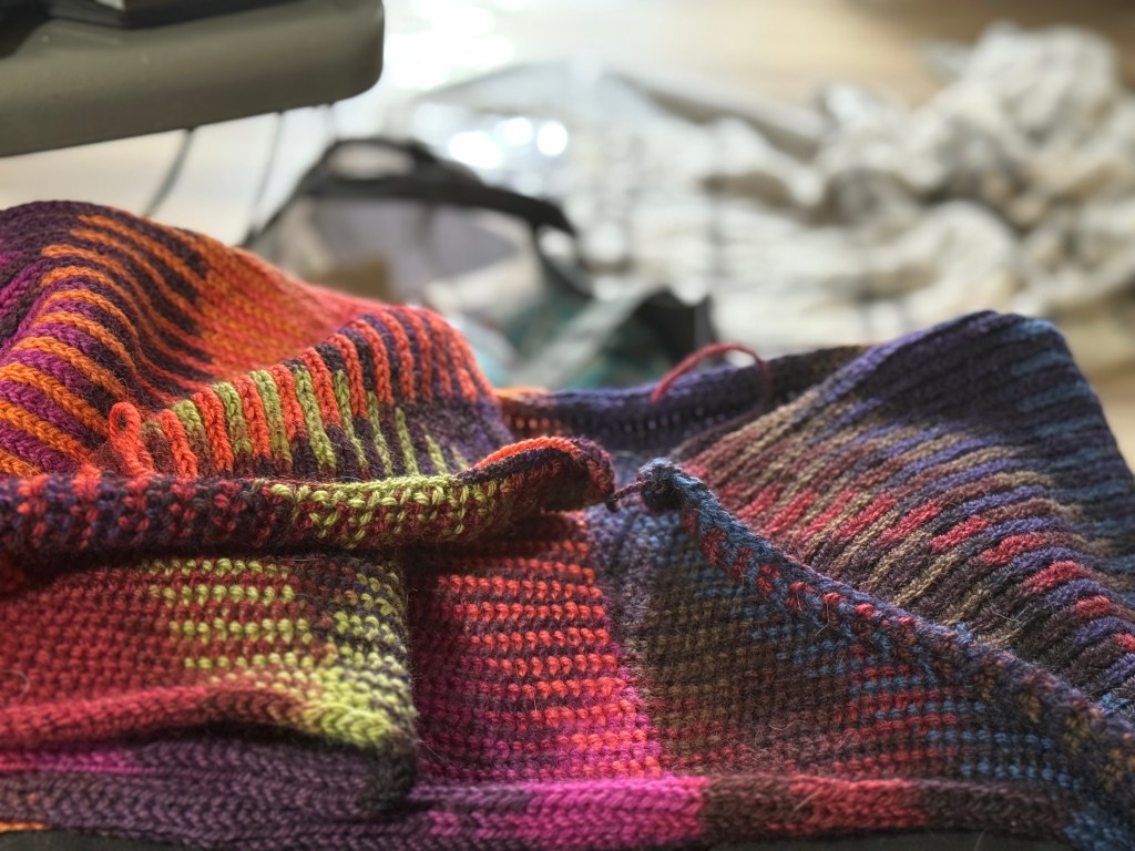 A close-up of a knitted Sweetspot strip in progress, showing folded sections of textured stitches and striking colour shifts. The variegated yarn reveals oranges, greens, purples, and blues in crisp stripes and pooling blocks.