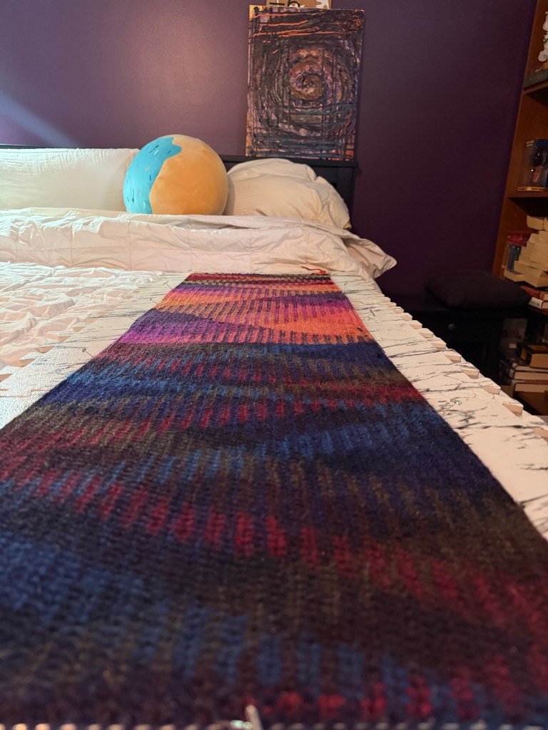 A hand-knit strip is pinned flat for blocking on a bed. The gradient pattern of the strip features rich purples, deep blues, and hints of red, transitioning into vibrant pink and orange tones toward the top. The scarf is framed by marble-print blocking mats.