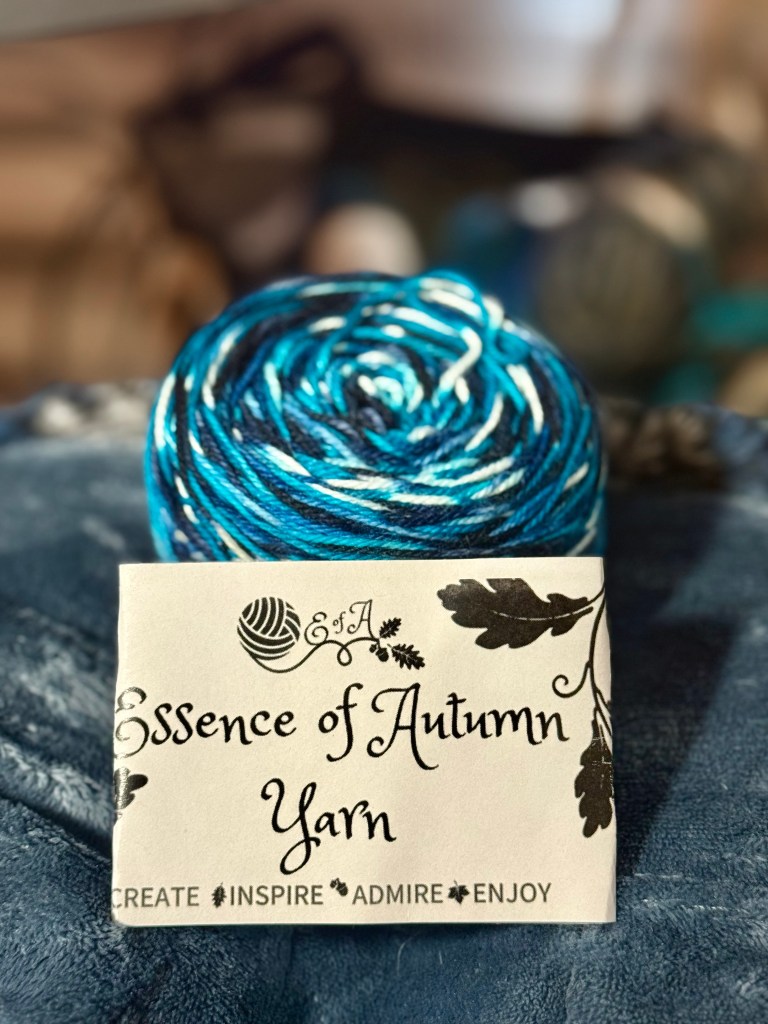 A close-up of a wound yarn cake in rich shades of blue, teal, black, and white, identified as “Sun Bleached Indigo” by Essence of Autumn Yarn. The cake sits on a soft navy blanket, with a label reading “Essence of Autumn Yarn” and the words “Create • Inspire • Admire • Enjoy” beneath stylized oak leaves.
