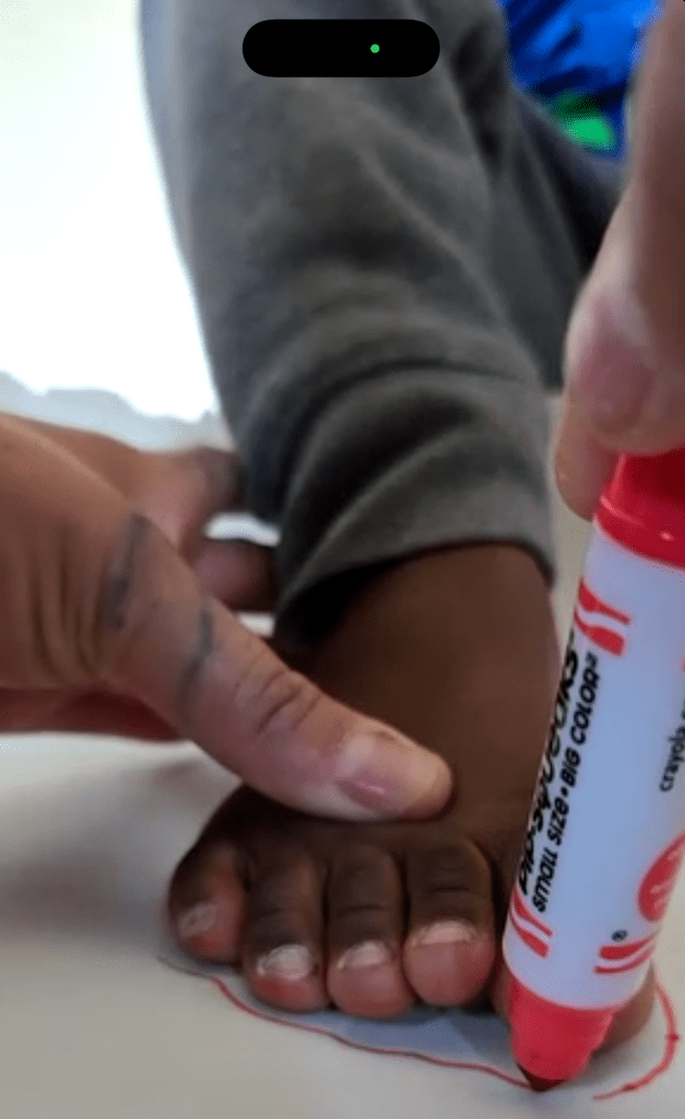 Close-up of a black child's foot being traced on white paper with a red marker. An adult’s hands gently hold the foot and marker in place as the outline is drawn. The image is a screen capture of a FaceTime call; the image of a caucasian woman, smiling widely, wearing a purple t-shirt, glasses pushed up on her head, in front of a bright yellow wall is in the bottom right corner.