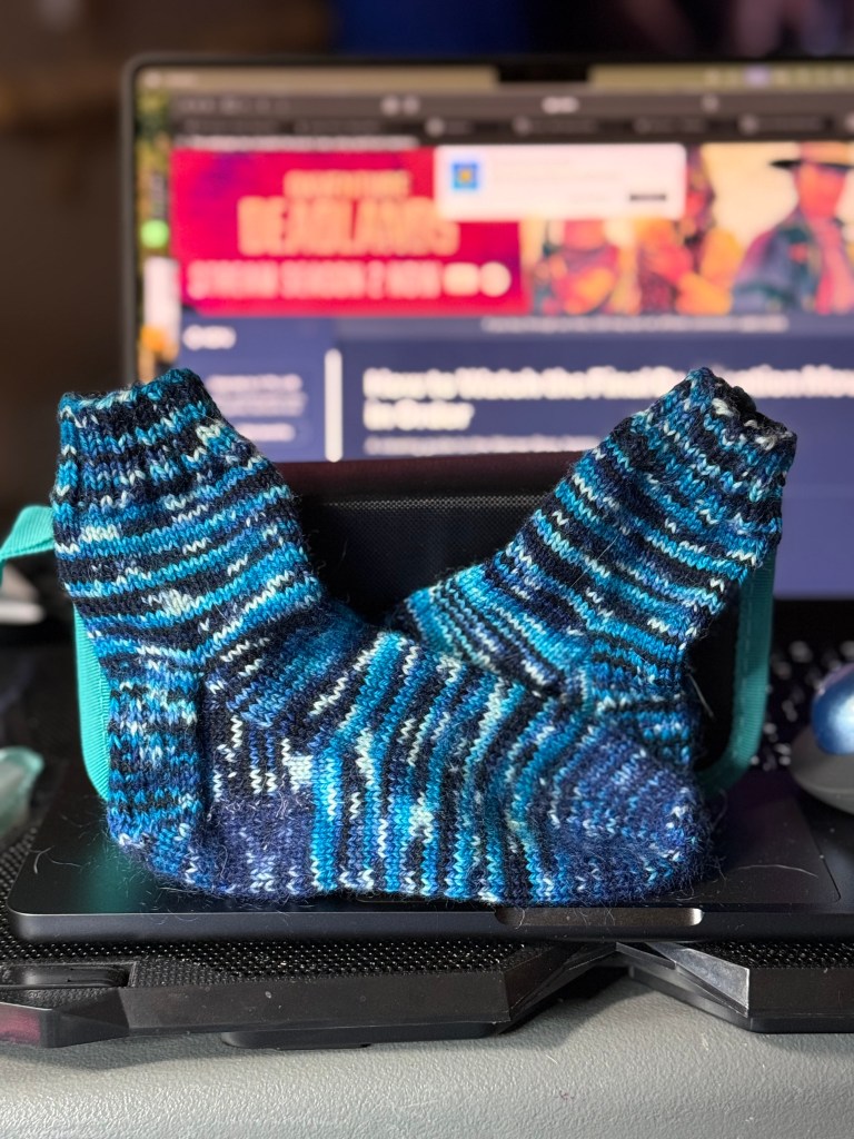 A pair of small, hand-knit socks in variegated shades of blue, black, and white are propped up against a teal case on a laptop keyboard. The background screen shows a paused video or webpage with vibrant red and blue hues, creating a cozy but tech-savvy workspace vibe.