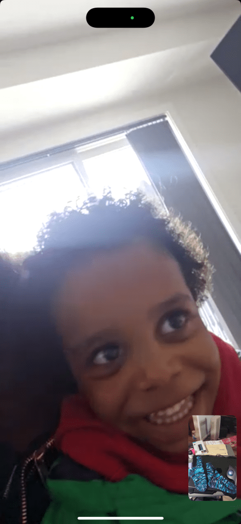 A smiling black child wearing a red and green jacket beams joyfully during a FaceTime call. The screen shows a small preview window of the caller, displaying a close-up of the finished pair of hand-knit socks. The sunlight from the window behind the child creates a warm, backlit glow..