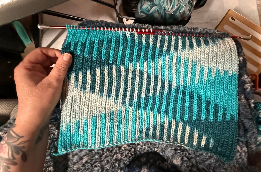 A close-up of a hand holding a piece of Knit One Below (K1B) fabric in variegated yarn featuring shades of teal, turquoise, and creamy white. The stitch pattern creates a vertical ribbed texture with dramatic colour pooling, forming wave-like diagonal patterns. The swatch is still on the needles and rests on a plush grey background. A tattooed forearm holds the edge of the work.