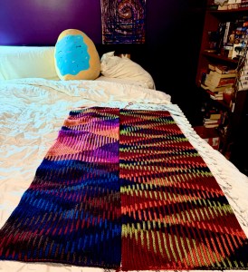 A vibrant, hand-knit panel made with the Knit One Below (K1B) stitch lies flat on a white bedspread. The piece is divided into two vertical sections, each showcasing dramatic planned pooling in saturated, high-contrast variegated yarn. The left half features a blend of pink, purple, peach, and navy, forming diagonal bands. The right half displays fiery reds, greens, and oranges in more vertical, ladder-like pooling. In the background, a plush donut pillow rests on white pillows against a deep purple wall, with a bookshelf and textured abstract artwork completing the cozy, artistic setting.