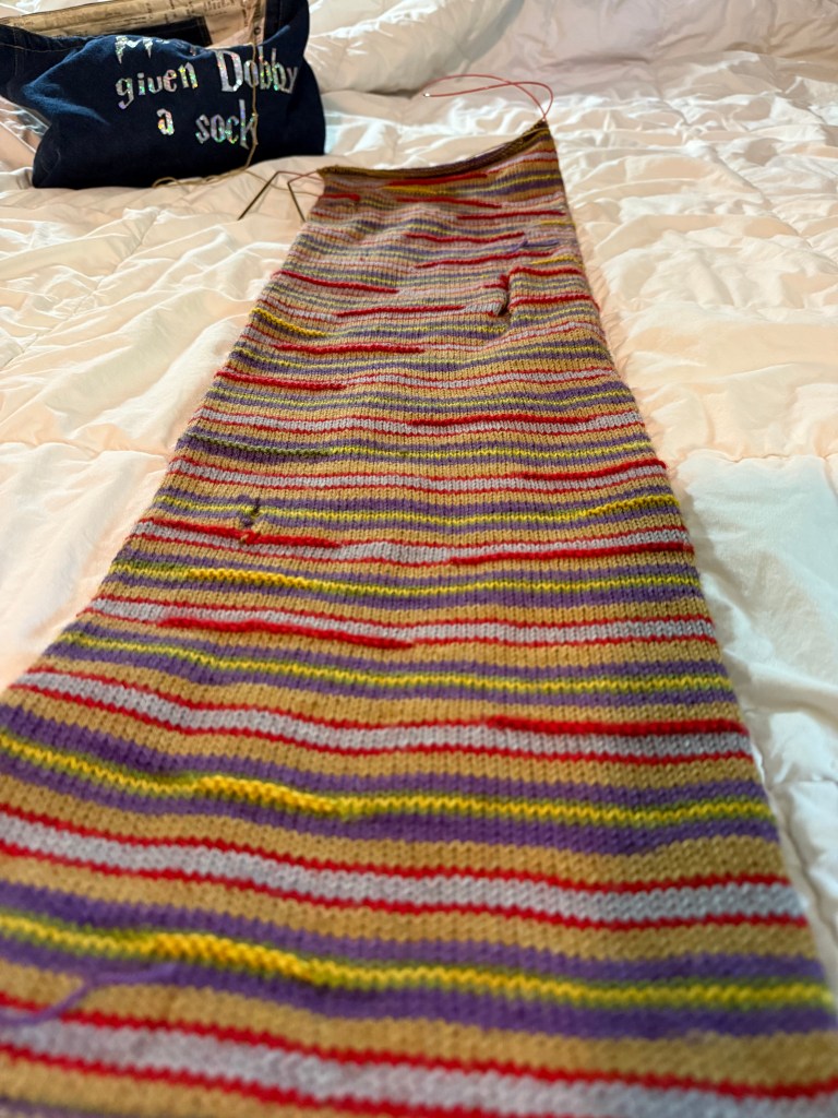 A striped scarf-in-progress in red, yellow, purple, grey, and tan lies on a white comforter with a dark blue project bag in the background that reads "Dobby is Free." Circular needles are visible at the top of the work.