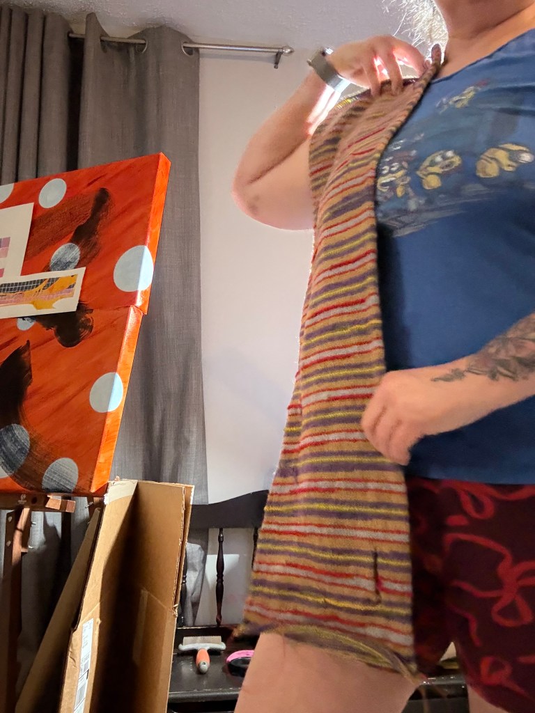 A female (partially visible) holding the scarf up to their chest to show its current length. She is wearing a faded mid-blue t-shirt with Minions on the chest, and a pair of Woxer ballers in a dark red with bright red bows printed on them. An abstract painting and art supplies are visible in the background.