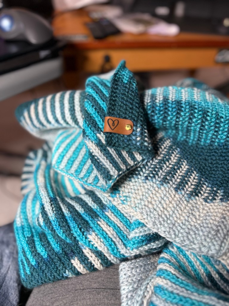 Another angle of the same knitted blanket, highlighting the reverse side of the custom leather label. This side features a simple stamped heart icon. The intricate striping of the Sweetspot pattern is visible in more detail, showcasing tonal transitions from dark teal to icy blue and soft grey. The blanket is draped casually across a lap and a grey cushion, with a desk and tabletop blurred in the background