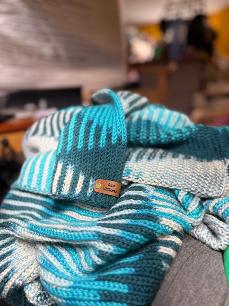 A close-up of a knitted blanket in rich shades of teal, turquoise, cream, and navy, arranged in rhythmic stripes using the Sweetspot pattern. A light brown leather tag is snapped onto the edge of the fabric, engraved with the name “Ace Williams” in black. The background is softly blurred, showing a cozy, lived-in room with warm lighting and a hint of wood furniture.