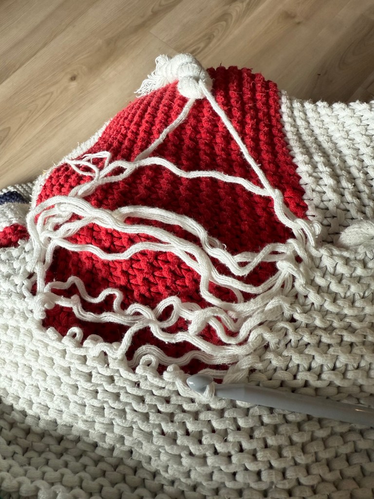 A close-up of a red and white section of a knit Hudson’s Bay blanket showing a large section of dropped white stitches over the red knit base. Loose strands are visible across the open gap, and a crochet hook is inserted at the edge, ready for repair.
