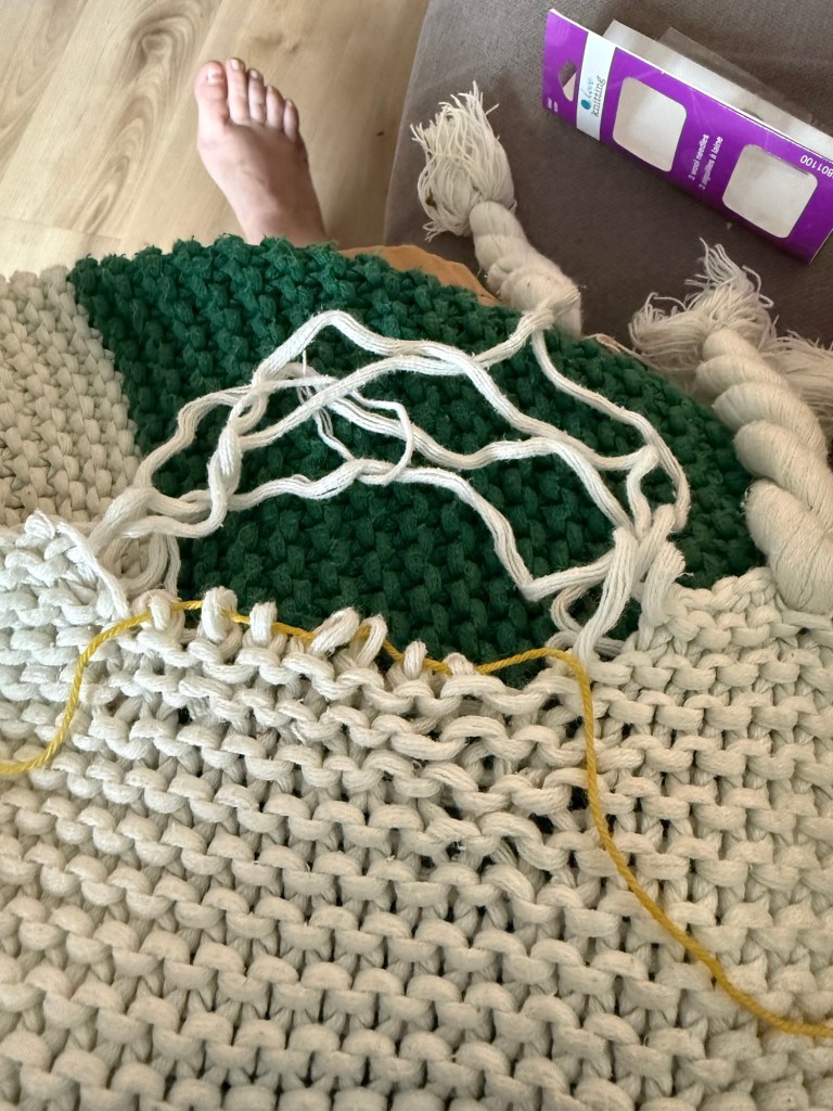 A knit Hudson’s Bay blanket with dropped white stitches. A yellow yarn lifeline is threaded through live stitches at the edge. Some loops are pulled up, and repair is in progress. A bare foot and a purple needle pack are in the background