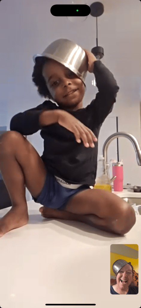 A young black child, smiling playfully during a FaceTime call, balances the same metal bowl on their head like a hat. The adult in the corner video frame is laughing and also wearing a bowl.