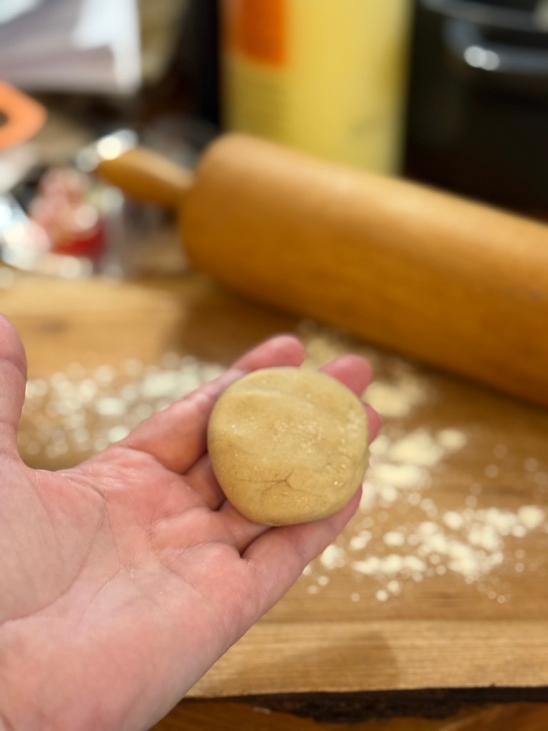 A hand holds a small, round, smooth ball of roti dough. The wooden surface beneath is lightly dusted with flour and a rolling pin sits nearby.