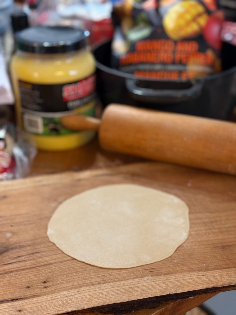 A single roti rolled out into a thin circle sits on a wooden board, ready to be cooked. A rolling pin and container of ghee are visible in the background.