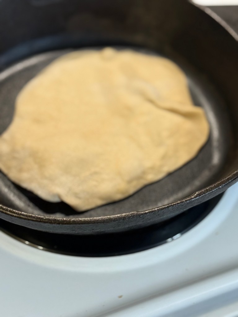An uncooked roti is cooking in a cast iron skillet. The dough is just beginning to change color as it heats.