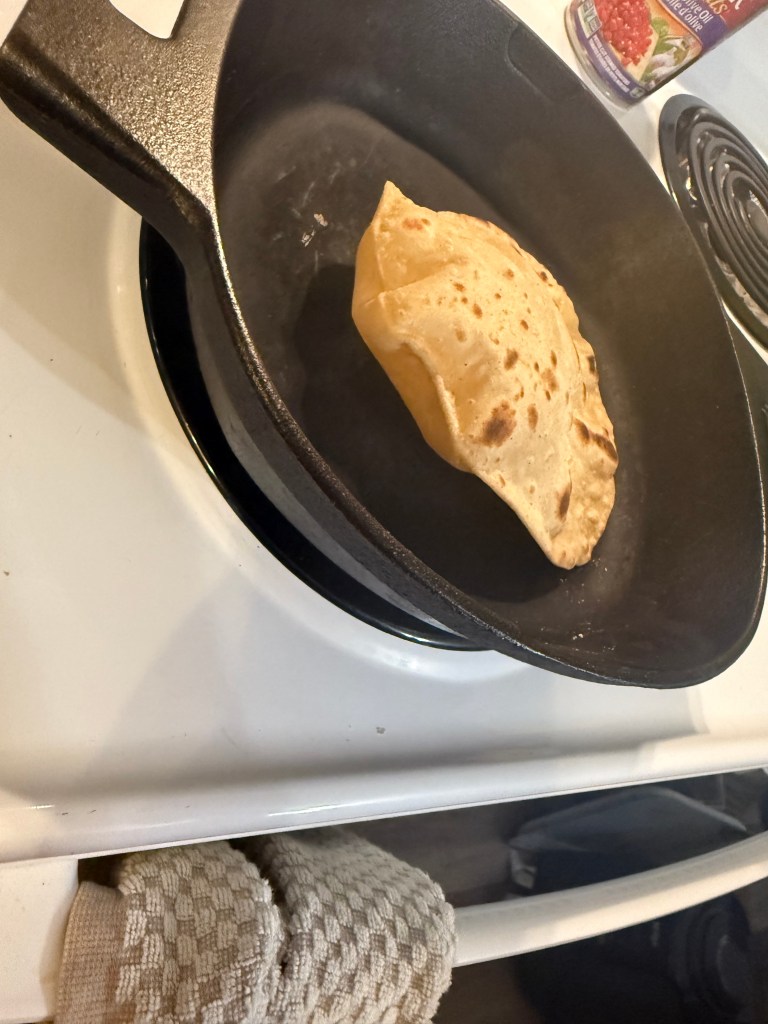 The roti has puffed dramatically in the skillet, forming a balloon-like shape with charred patches and perfect browning.
