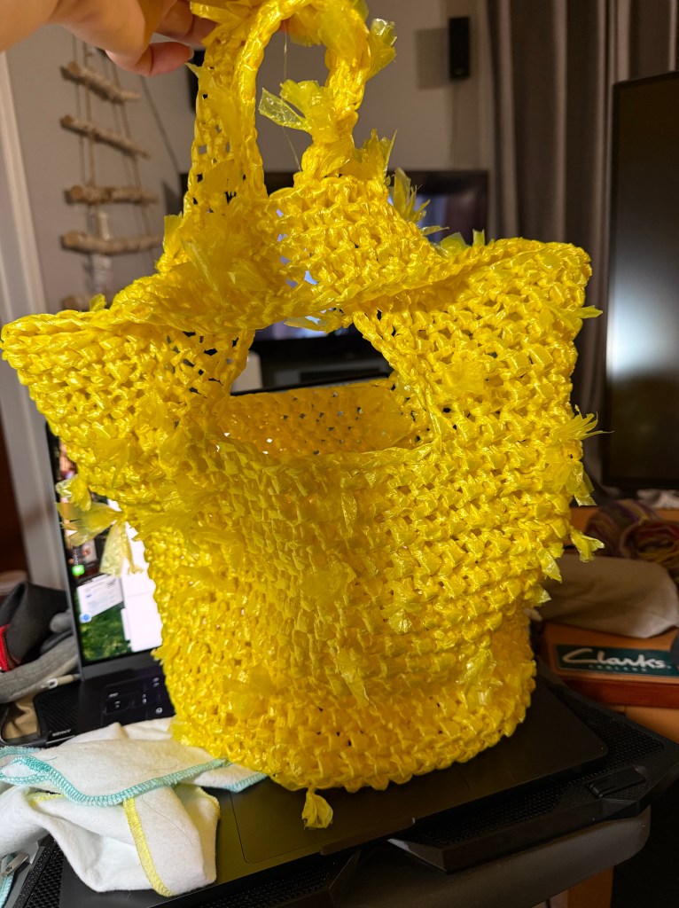 The same yellow crocheted plarn tote shown from the side, with its longer handle looped through the shorter one. This serves as a closure of sorts to the bag while still providing a sturdy single handle. The bag has a flexible, basket-like shape.