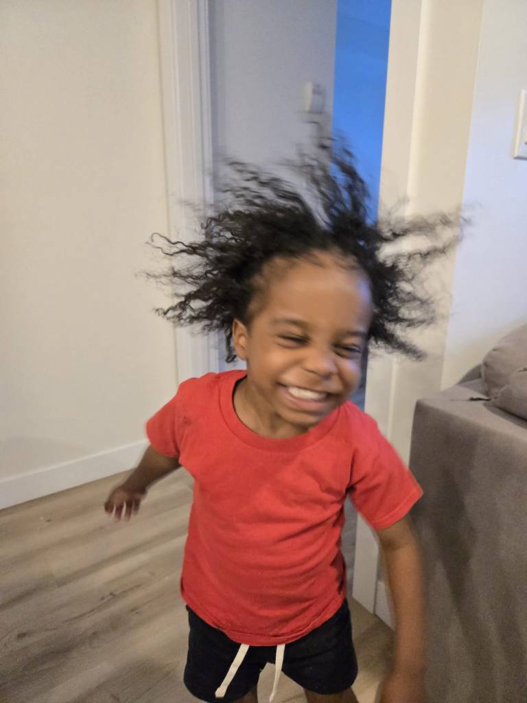 A 4 year old black boy wearing a red t-shirt and black shorts with white ties hanging down is captured mid-run, a priceless grin on his face. His hair is standing out all over his head as if he is conducting electricity. His hair has just been unbraided and he's delighted!!