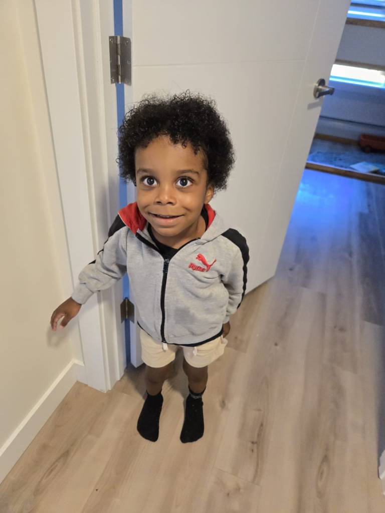 a 4-year old black boy is looking up at the photographer. He is wearing black ankle socks, tan shorts with the ends of white ties just showing and a grey Puma zip up sweater with black and red accents. His hair is naturally coiffed, some gel taming the curls. His wide eyes and genuine smile make him look so sweet.