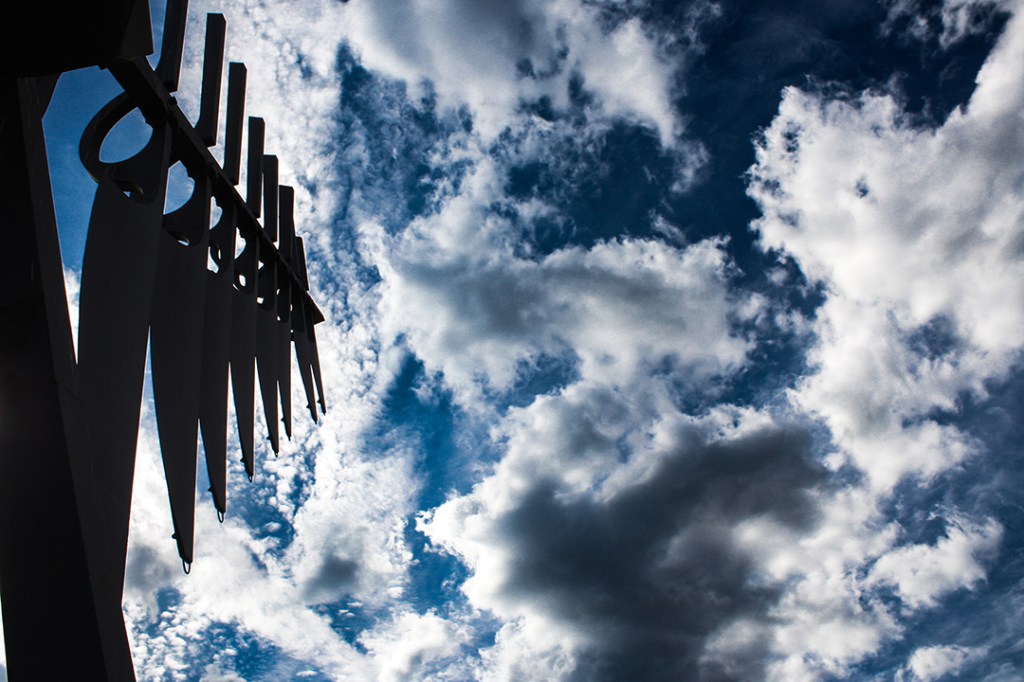 The Spirit Catcher sculpture in Barrie, Ontario. One wing of the sculpture is captured, darkly silhouetted on the left thrid of the image against a dramatic sky filled with billowing clouds, evoking the sense that a storm has been summoned.